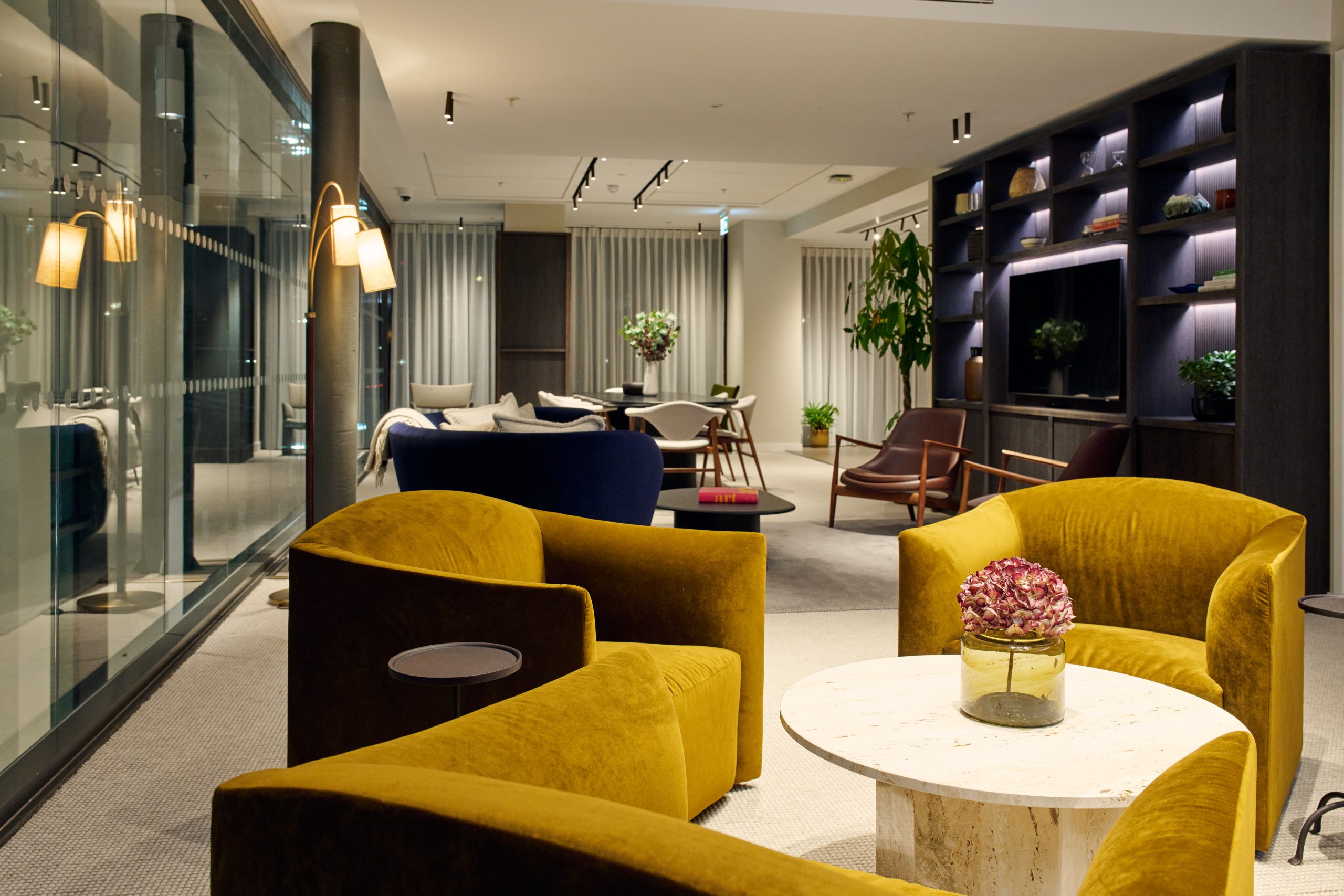 Communal lounge area showing three yellow velvet armchairs, a book case with TV and a table and chairs in the background