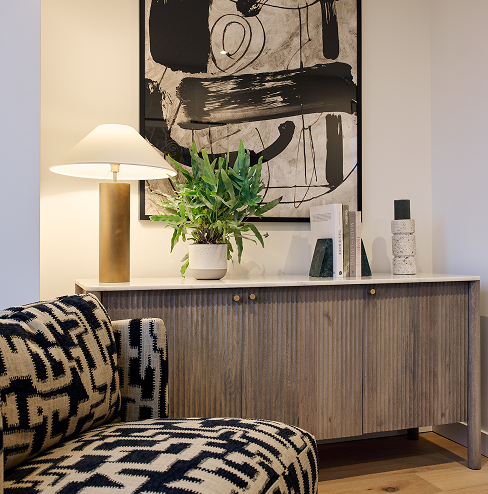 New apartment living area detail showing armchair in geometric print and side table with feature table lamp, plant and books on top and a piece of art on the wall behind