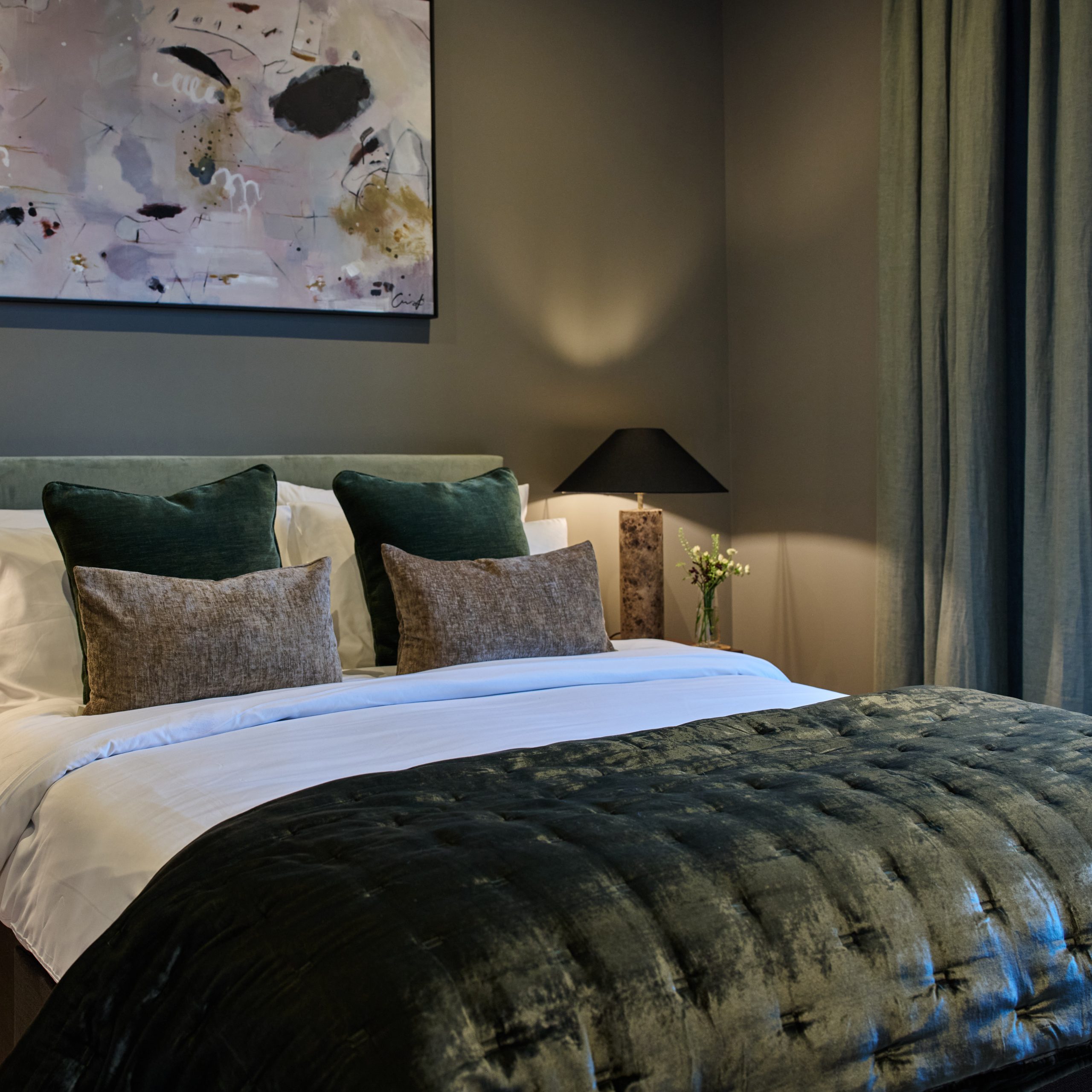 New apartment master bedroom at dusk showing a double bed with dark green velvet throw and cushions and a side table with table lamp
