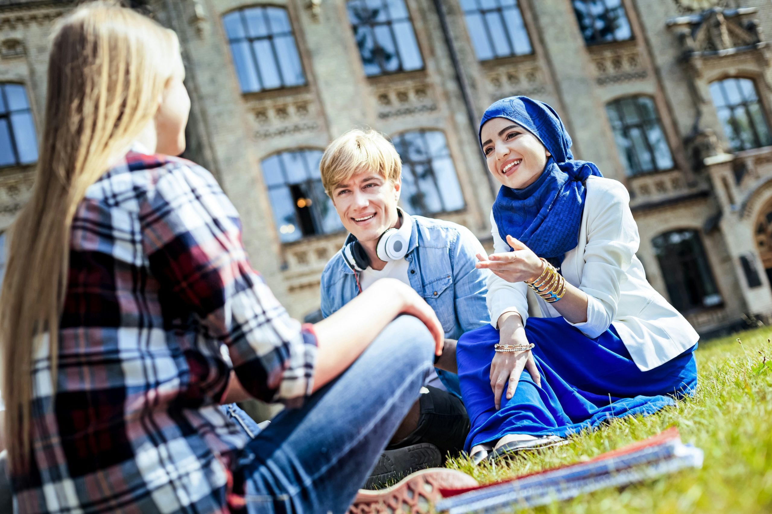 Three young people sitting on the grass talking in front of a traditional brick building