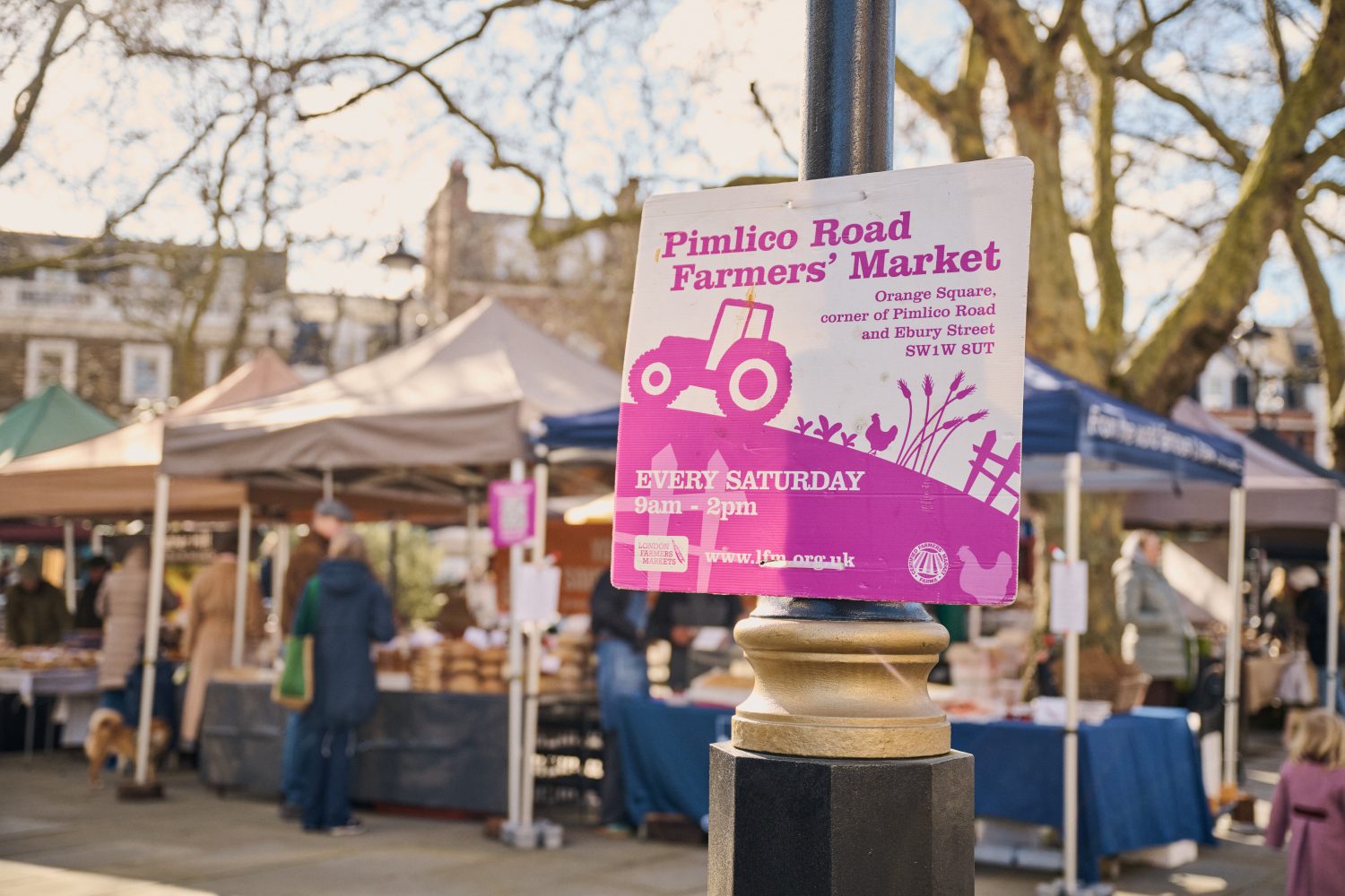 A lamppost sign advertising the Pimlico Road Farmers' Market with stalls in the background showing the market in full swing