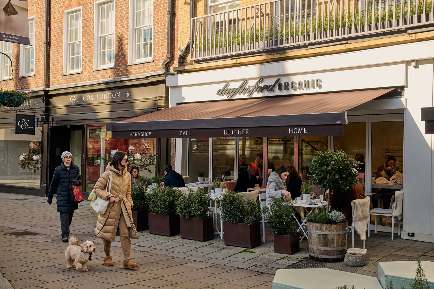People walking past an organic grocery looking across at people sitting at outside tables enjoying a cup of coffee.