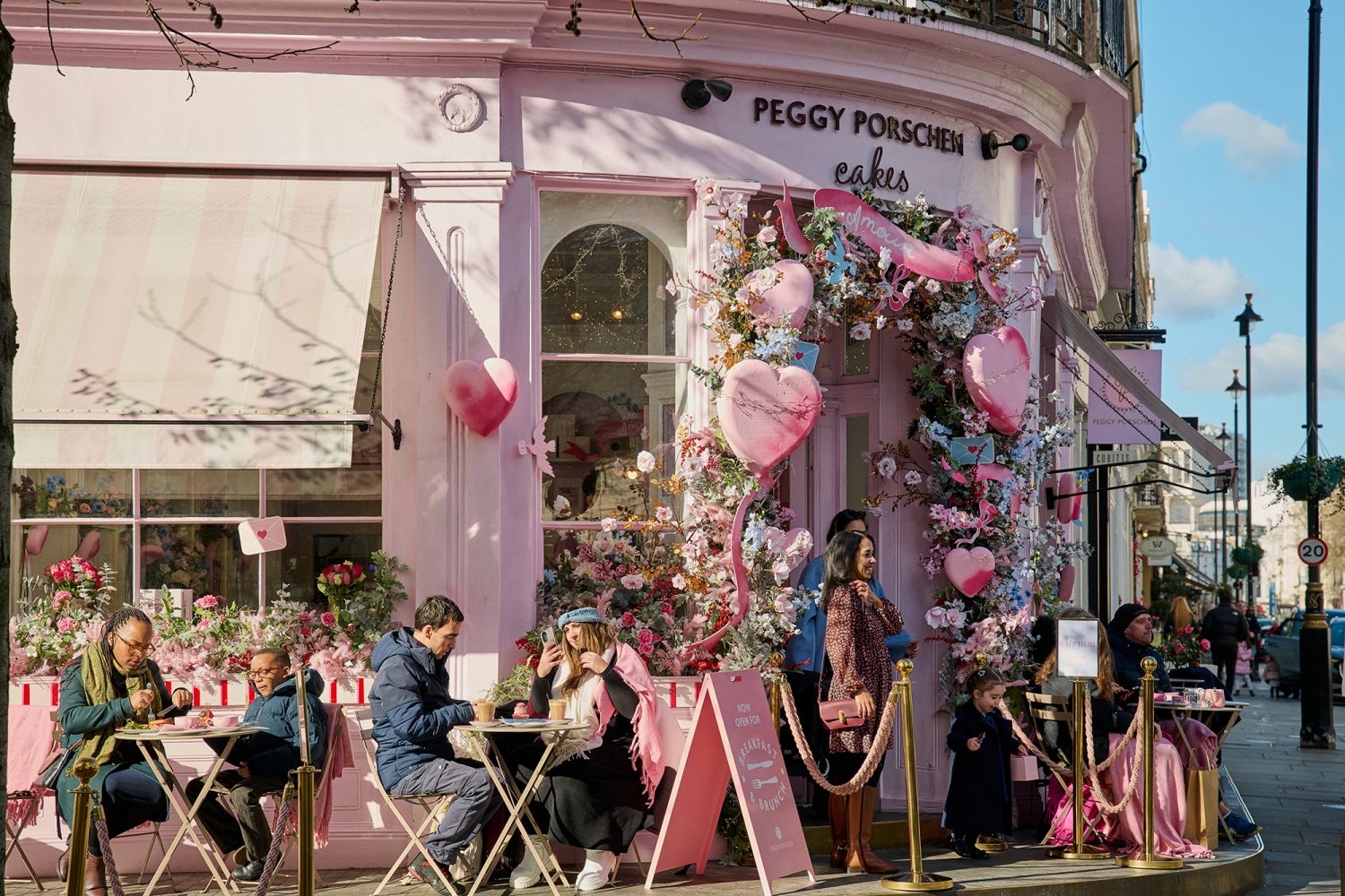 Pretty pink corner cafe with people seated outside eating cakes and pastries with a doorway decorated with flowers and balloons