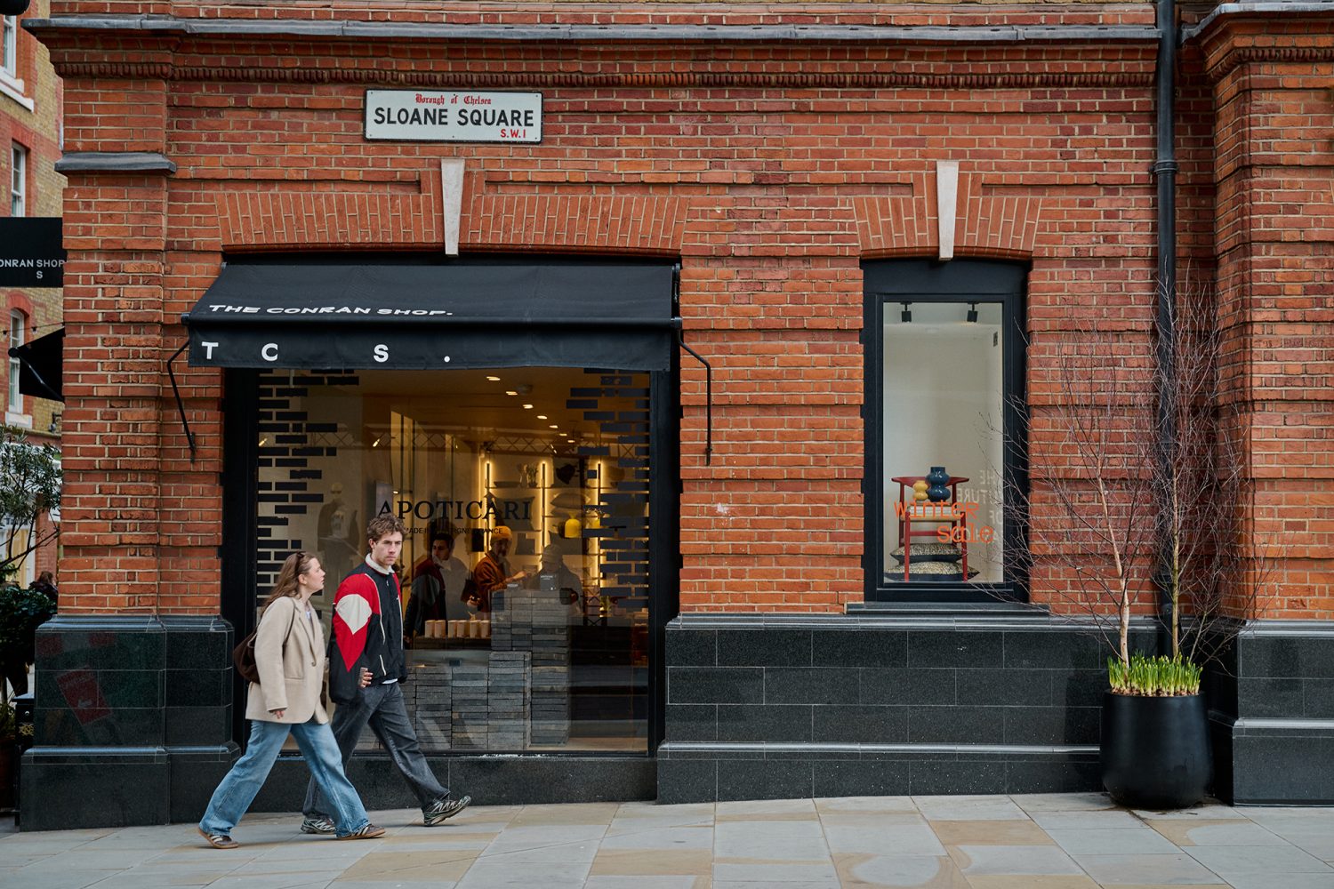 A couple walking past a brick fronted building with a Sloane Square street sign mounted on the building