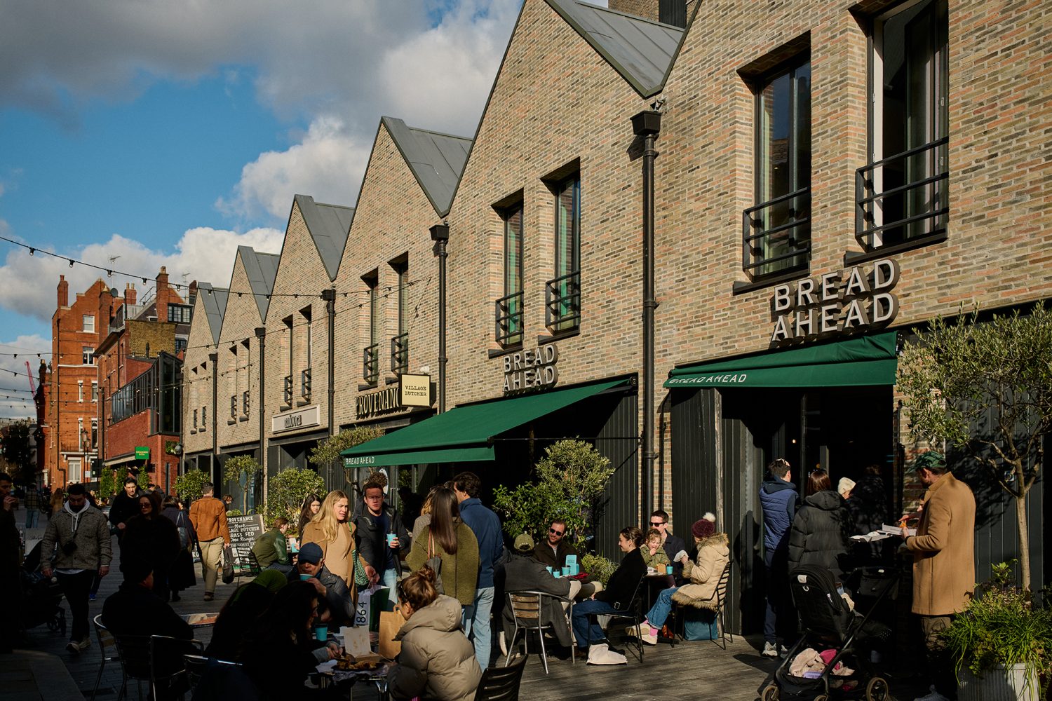 People gathered outside a bakery in an attractive street featuring a row of brick-fronted shops