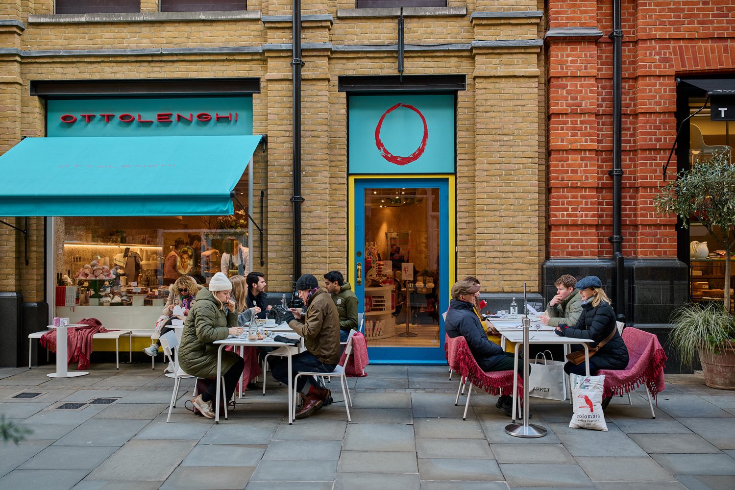 People enjoying coffees outside Ottolenghi deli. The chairs are bright pink and the awning outside the deli is turquoise