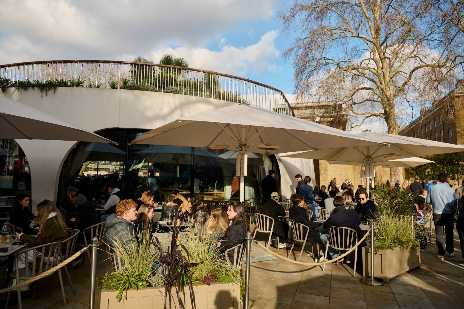 People eating in a busy outside seating area at a circular restaurant with large windows